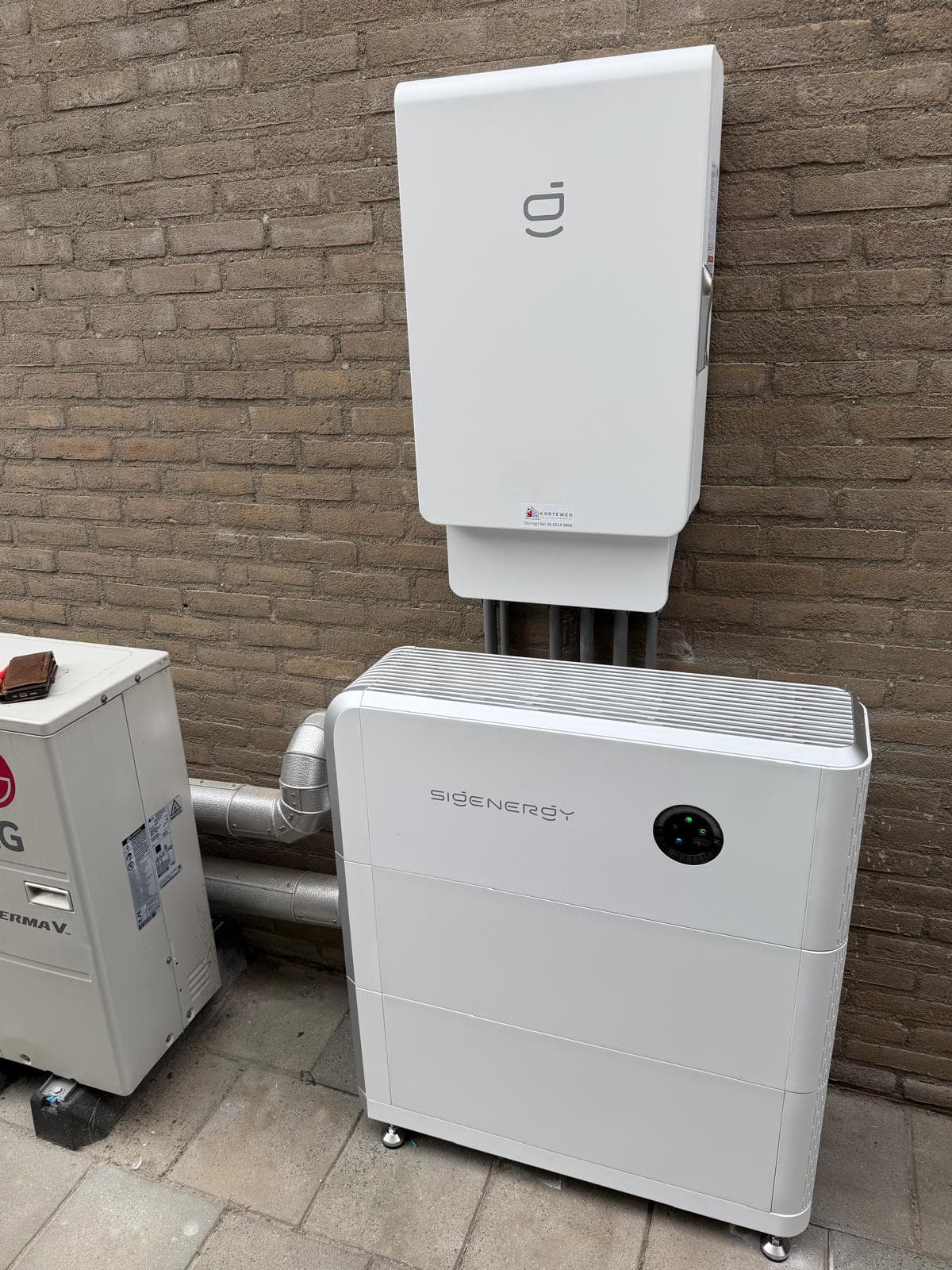 Sigenergy-thuisbatterij met omvormer erboven, geplaatst aan een bakstenen gevel